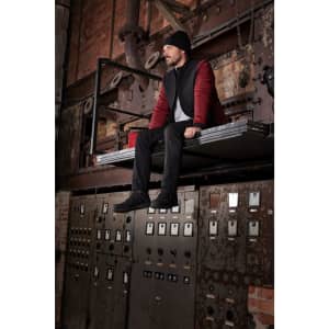 A men's padded hybrid jacket in red and black, worn by a person sitting on an industrial shelf. Features branding.