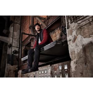 A man wearing a red and black padded hybrid jacket sits on a metal platform in an industrial setting.