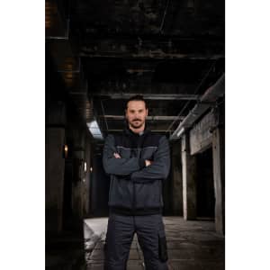 A man wearing a black and grey Hybrid Workwear Jacket stands confidently in an industrial setting.