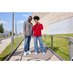 A red fleece jacket and a grey fleece jacket worn by a couple, standing on a pathway with greenery and a building in the background.