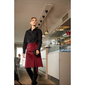 A woman wears a burgundy apron with a black shirt, standing in a stylish indoor setting with modern lighting.