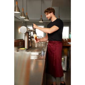 A man wearing a burgundy apron pours a drink from a tap in a bar setting. The image features modern pendant lights.