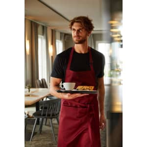 A maroon apron with bib displayed on a model holding a tray with a coffee and food in a restaurant setting.