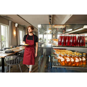 A man wearing a burgundy apron with a bib carries a plate in a modern dining setting, surrounded by drinks in glass containers.