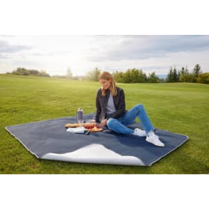 A picnic blanket in navy and white, laid out on grass with a woman sitting, surrounded by snacks and a drink bottle.