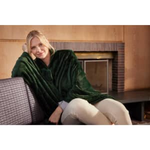 A green microfibre fleece blanket XL draped over a seated woman on a couch, with a fireplace in the background.