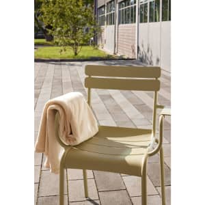 A beige microfibre fleece blanket draped over a light-green chair outdoors, with a paved pathway and greenery in the background.