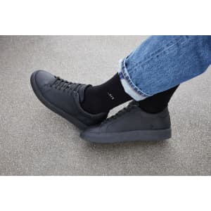 Black sports socks worn with black shoes and blue jeans on a grey surface. The socks have a logo.