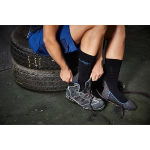 A person is putting on grey shoes while wearing black and grey socks, seated on tyres in a work environment.