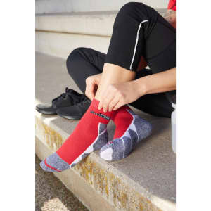 Sport socks in red with grey accents, featuring a logo, being worn by a person seated on a step.