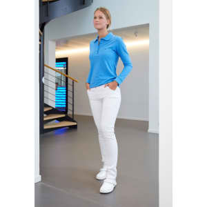 A model wearing white stretch pants and a blue long-sleeve polo top stands in a modern interior setting.