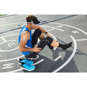 A man in a blue running tank and black leggings stretches on a sports track, wearing a visor and sneakers.