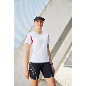 A woman wearing a white T-shirt with red accents and black shorts, standing outdoors near a structural backdrop.