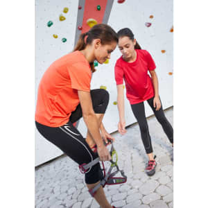 Two girls in vibrant orange and red T-shirts are preparing for a climbing activity at a rock wall.