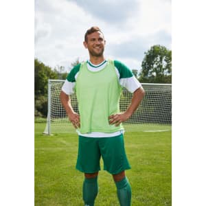 A young man stands confidently on a football pitch, wearing a bright green training bib, green shorts, and green socks.
