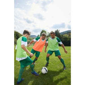 Three players in green and orange training vests engage with a soccer ball on a grassy field under a cloudy sky.