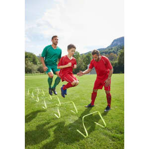 A training scene featuring two adult players in green and red shirts, coaching a young boy in a red shirt, jumping over hurdles.