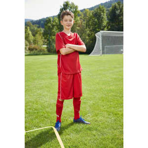 A young boy stands confidently in a red football kit on a grassy field, with goalposts in the background. The outfit has a logo.