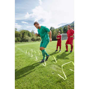 A man in green sports shorts and shirt performs agility drills over hurdles on a grass field, with two players in red watching.