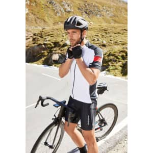 A man in a black and white cycling jersey adjusts his gloves while standing next to a bicycle on a mountain road.