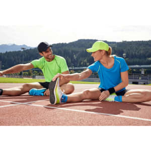 A pair of athletes stretch on a track, wearing vibrant green and blue sports T-shirts and accessories.