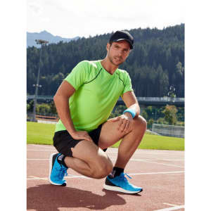 A man in a bright green sports T-shirt and black shorts kneels on a running track, wearing blue athletic shoes and a cap.
