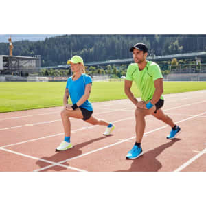 A man in a green sports t-shirt and a woman in a blue sports t-shirt perform lunges on a running track. Both wear caps.