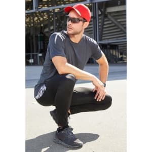 A man wearing a grey T-shirt and black leggings, complemented by a red cap and sunglasses, poses outdoors.