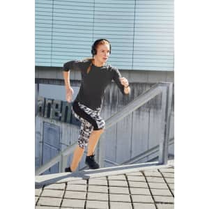 A woman in a long-sleeve grey sports shirt and patterned leggings running up outdoor stairs, wearing headphones.