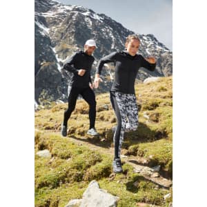 A black ladies sports zip hoodie is worn by a woman running through a mountainous landscape, alongside a man in a cap.
