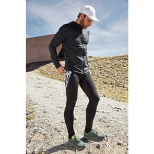 A man in a black zip hoodie and black leggings stands outdoors, wearing a white cap and sporty shoes. The background features rocky terrain.