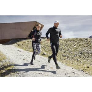 A man and woman jog outdoors wearing athletic outfits, including a black zip hoodie. The scenery is rugged and mountainous.