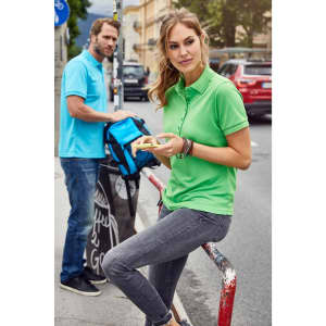 A light green ladies active polo shirt with a branding logo, worn by a woman sitting casually, paired with grey jeans.