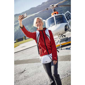 A woman in a red knitted fleece hoodie takes a selfie near a helicopter, wearing a backpack and waist bag.