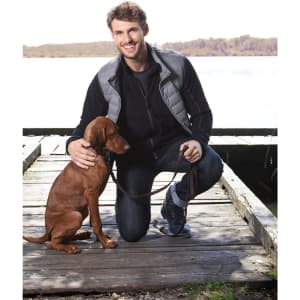 A man in a black fleece jacket kneels beside a brown dog on a wooden dock by a lake. The scene is outdoor and sunny.
