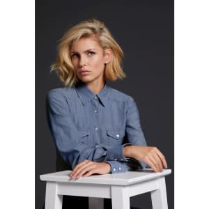 A denim blouse in light blue with button-up front and chest pockets, displayed by a model sitting at a white table.