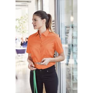 A woman wearing an orange short-sleeve button-up shirt stands in a bright indoor setting, holding a phone.