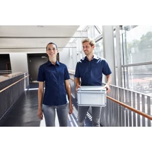 A navy ladies business button-up shirt is worn by a woman, accompanied by a man holding a container in a modern hallway.