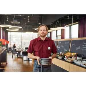 A man in a burgundy button-up shirt holds stacked plates in a modern dining area with large windows and a buffet setup.