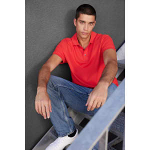 A red men's Pima polo shirt, modelled by a man sitting on stairs, wearing blue jeans and white sneakers.