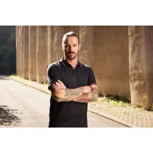 A man with tattoos is wearing a black polo shirt with a contrasting collar, standing against a textured background.
