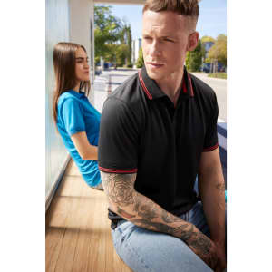 A black polo shirt with red accents is worn by a man sitting beside a woman in a blue polo shirt. Both are outdoors.