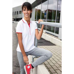 A woman wearing a white polo shirt with red accents, styled with grey jeans and red sneakers, stands outdoors.