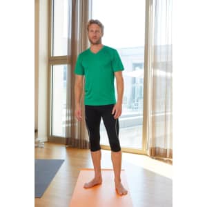 A man stands barefoot on a yoga mat wearing a green active T-shirt and black athletic leggings, with natural light streaming in.