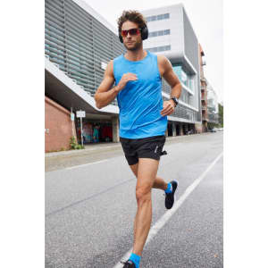A man in a blue active tank top and black shorts jogs along a city street, wearing sunglasses and headphones.