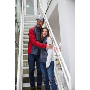 A man in a red and grey knitted hybrid jacket and a woman in a grey and white jacket stand on a staircase, smiling.