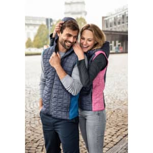 A knitted hybrid vest in navy with blue and pink accents, worn by a smiling couple outdoors. The vest has a hood and a logo.