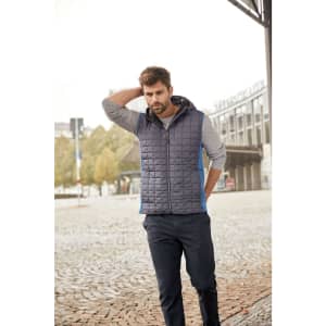 A man wears a navy and grey knitted hybrid vest with a hood, paired with a long-sleeve shirt and casual pants.