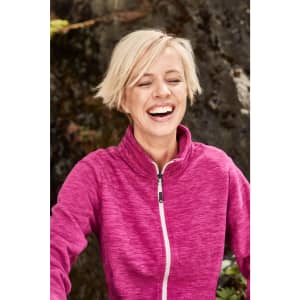 A woman wearing a pink polar fleece jacket, smiling against a natural backdrop. The jacket features a zip and a logo.