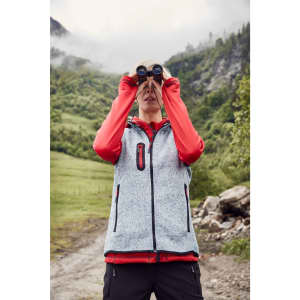 A woman wearing a grey knitted fleece vest with red accents, using binoculars outdoors in a hilly landscape.